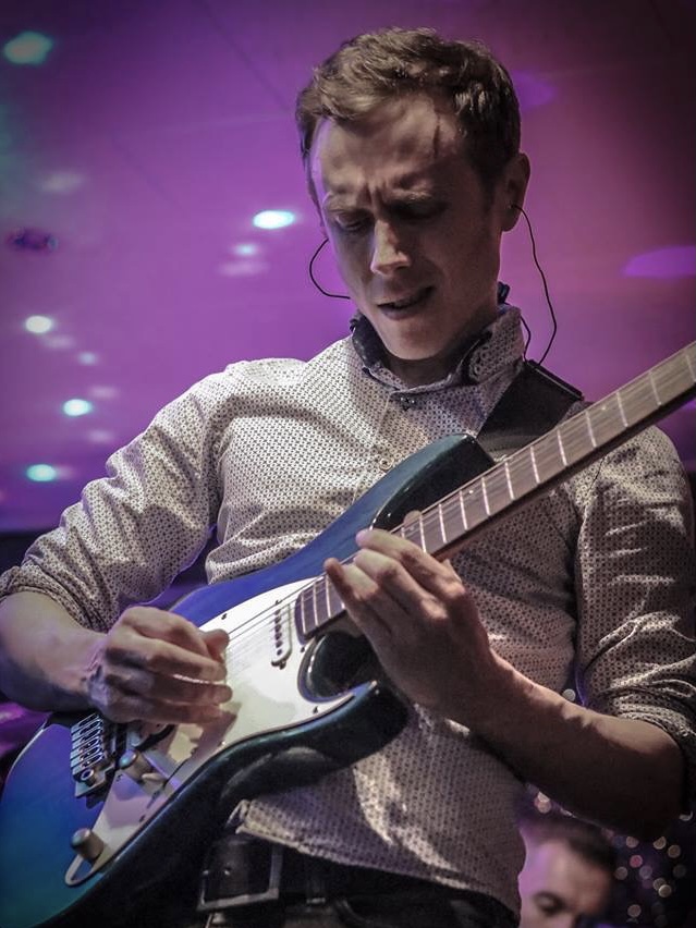 IndieFly guitarist Adam Smithson playing a blue guitar live. He is wearing a smart white shirt.