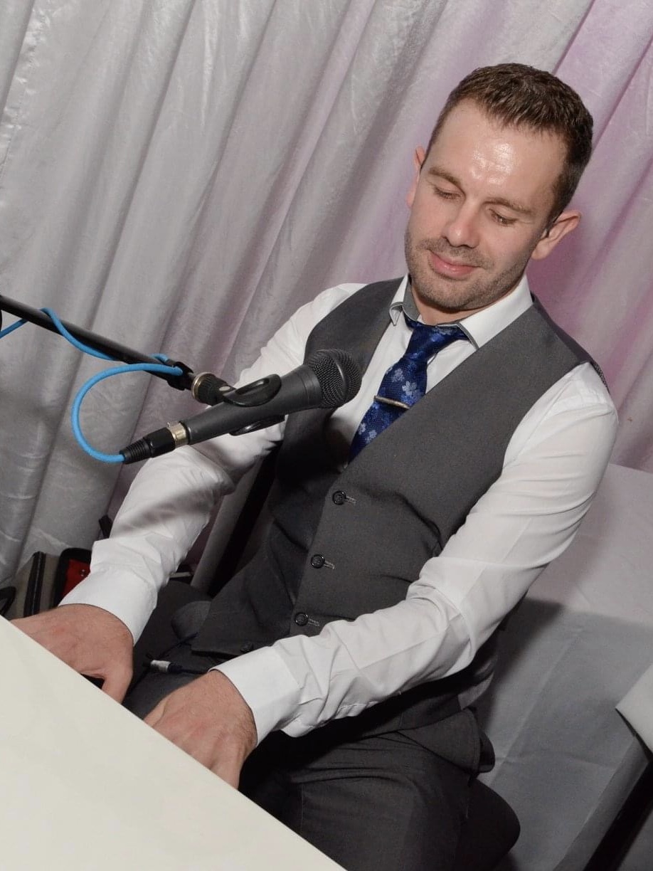IndieFly keyboardist and wedding planner Craig Smith wearing a suit and looking down at the keys of a white baby grand piano he is playing
