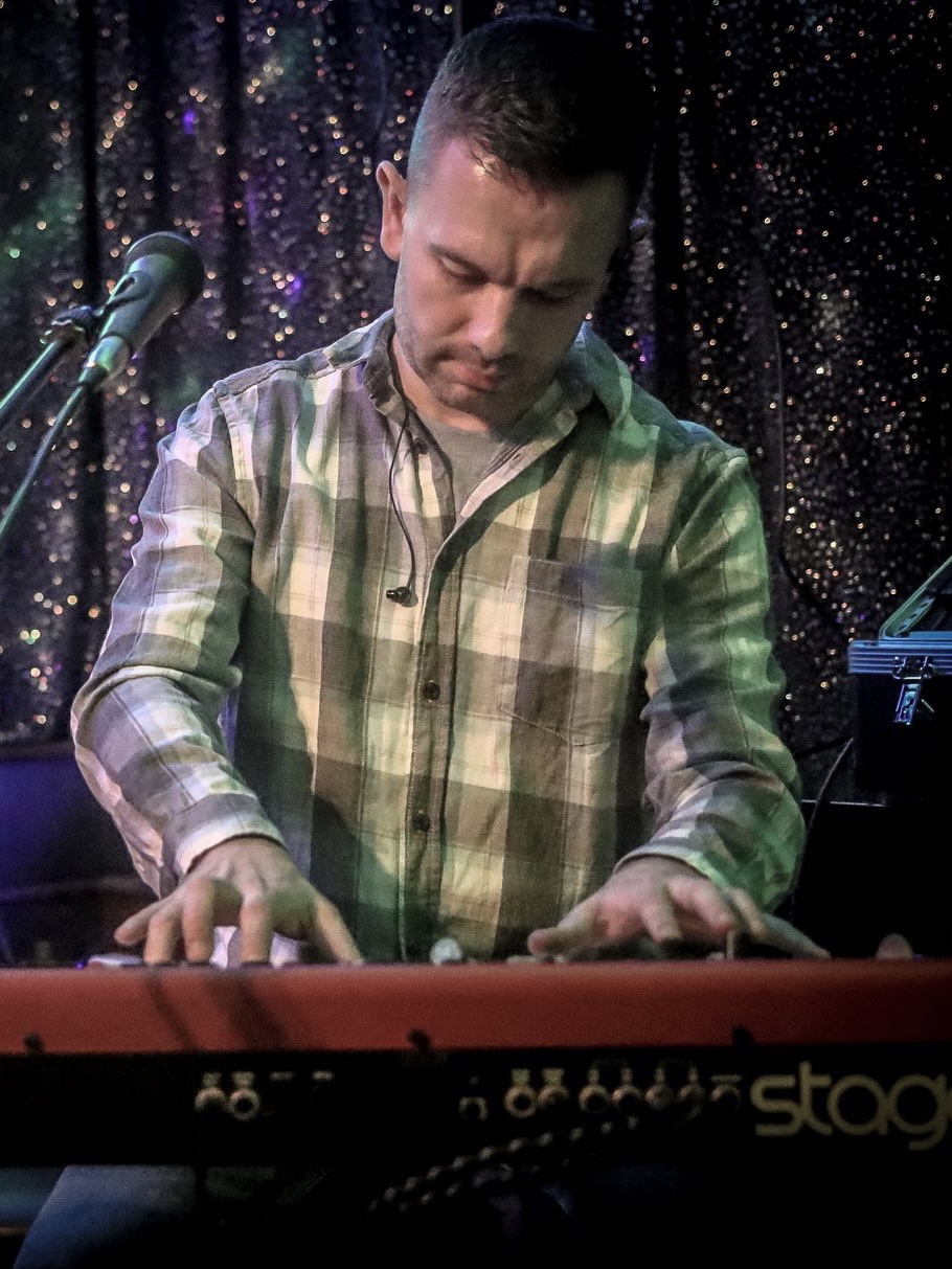 IndieFly keyboard and pianist player Craig Smith playing a red Nord stage piano and wearing a white shirt with a blue and grey check pattern